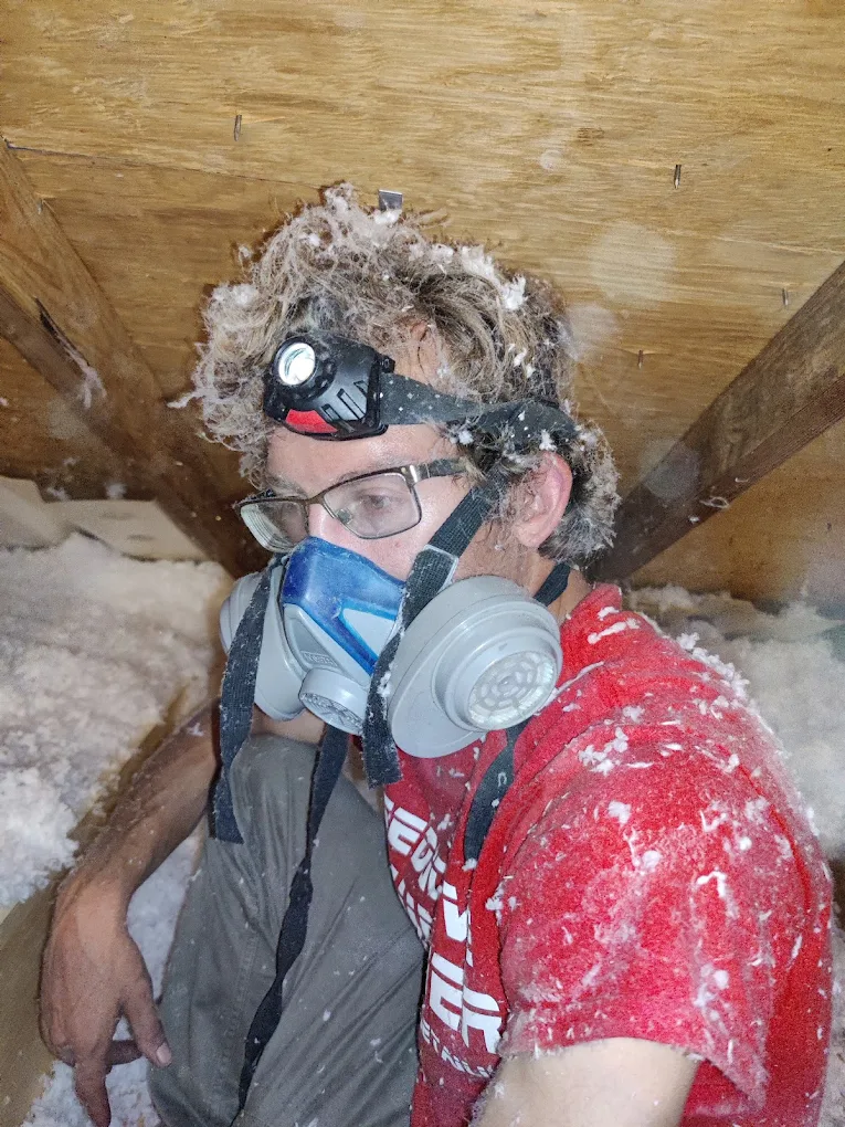 Joe in a respirator and headlamp covered in insulation inside a tight attic